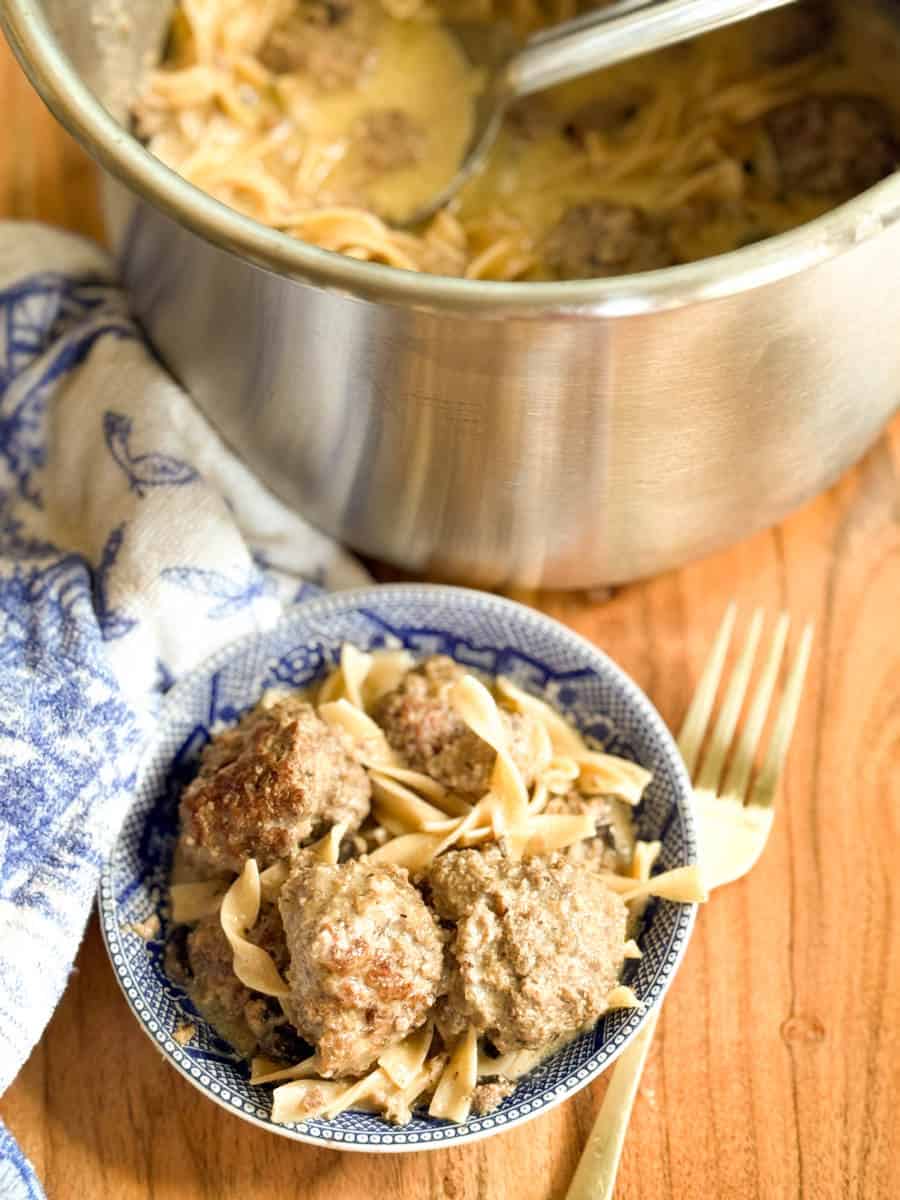 A small bowl filled with noodles and meatballs sits on a wooden table beside a fork. In the background, a pot with more noodles and meatballs and a blue-and-white towel are visible.