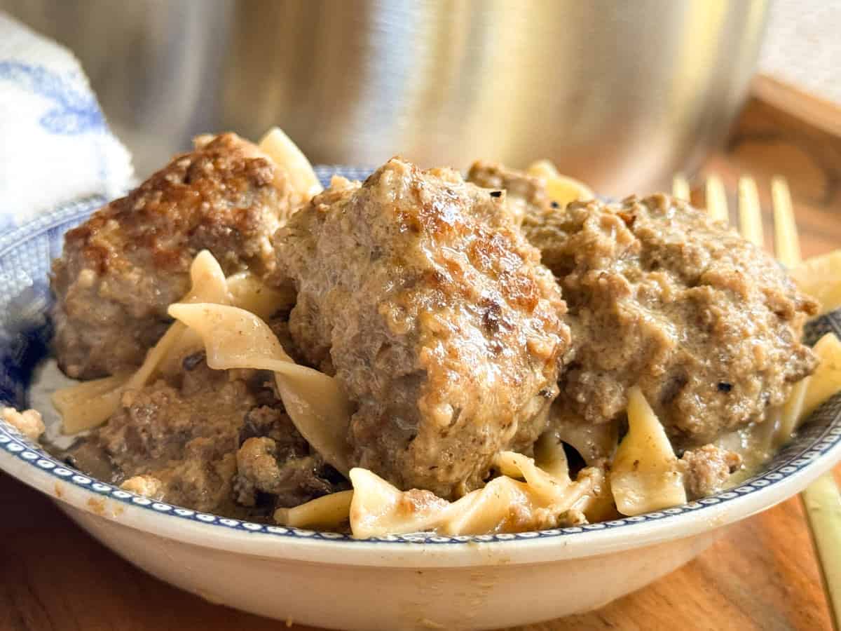 A close-up of a bowl containing browned meatballs served over egg noodles with a creamy sauce. A fork and part of a metal pot are visible in the background.