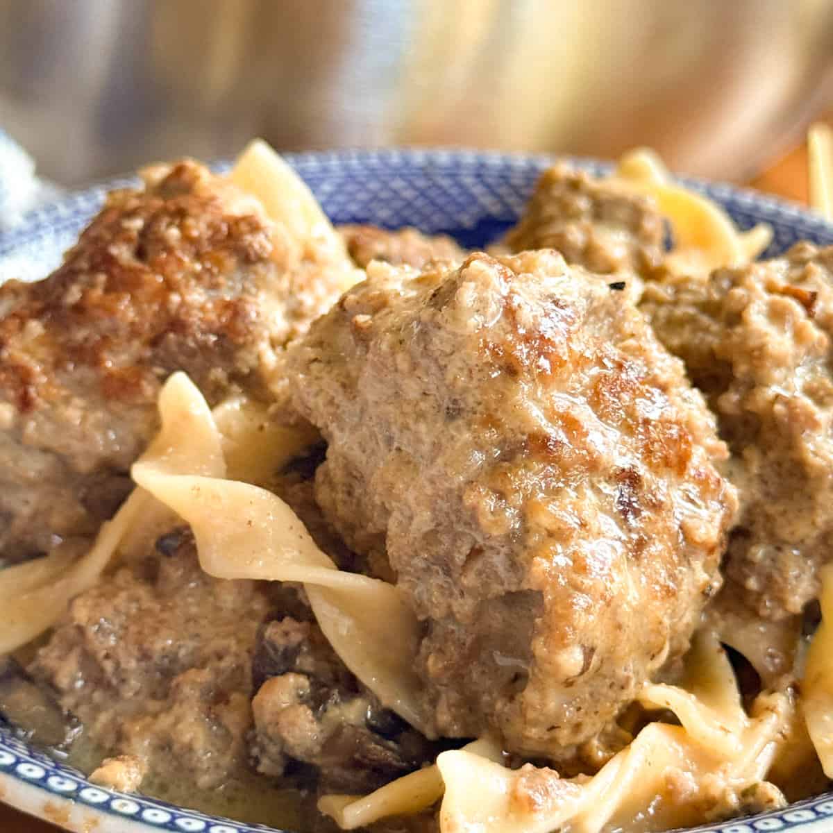 A blue patterned bowl filled with meatballs and wide egg noodles, covered in a creamy brown sauce. The dish appears freshly cooked with a rich, textured appearance.