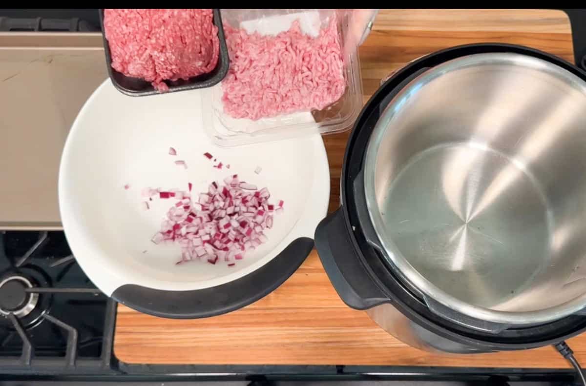 A mixing bowl with chopped red onion beside raw ground meat in plastic trays, next to an empty electric pressure cooker on a wooden countertop near a stovetop.