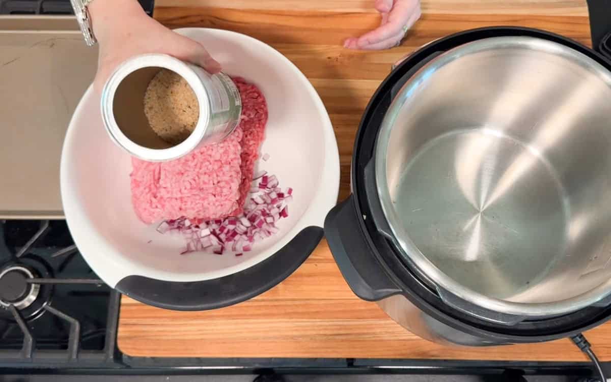A person adds seasoning from a can to a bowl containing ground meat and chopped red onions. An empty pressure cooker sits nearby on a wooden countertop next to a stovetop.