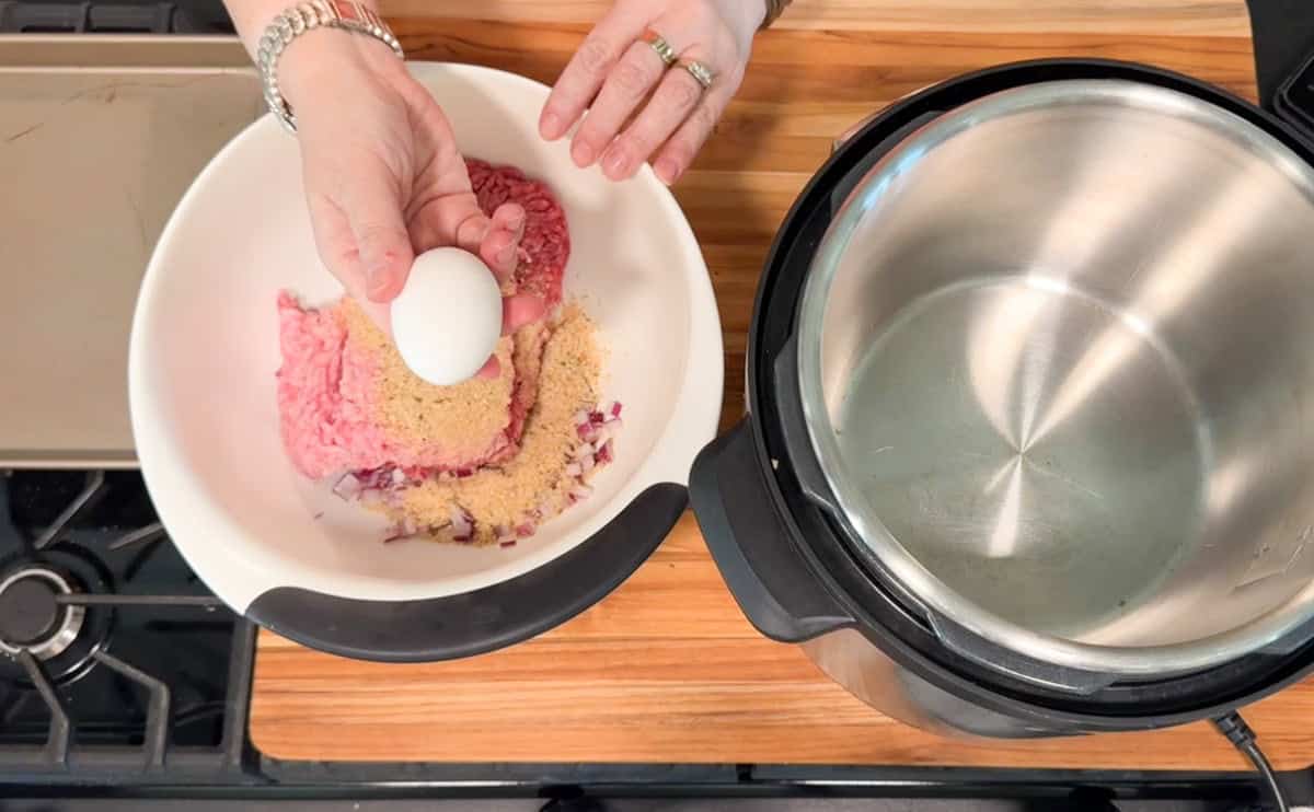 A person holds an egg above a white bowl containing ground meat, breadcrumbs, and chopped onions next to an open Instant Pot on a wooden countertop.
