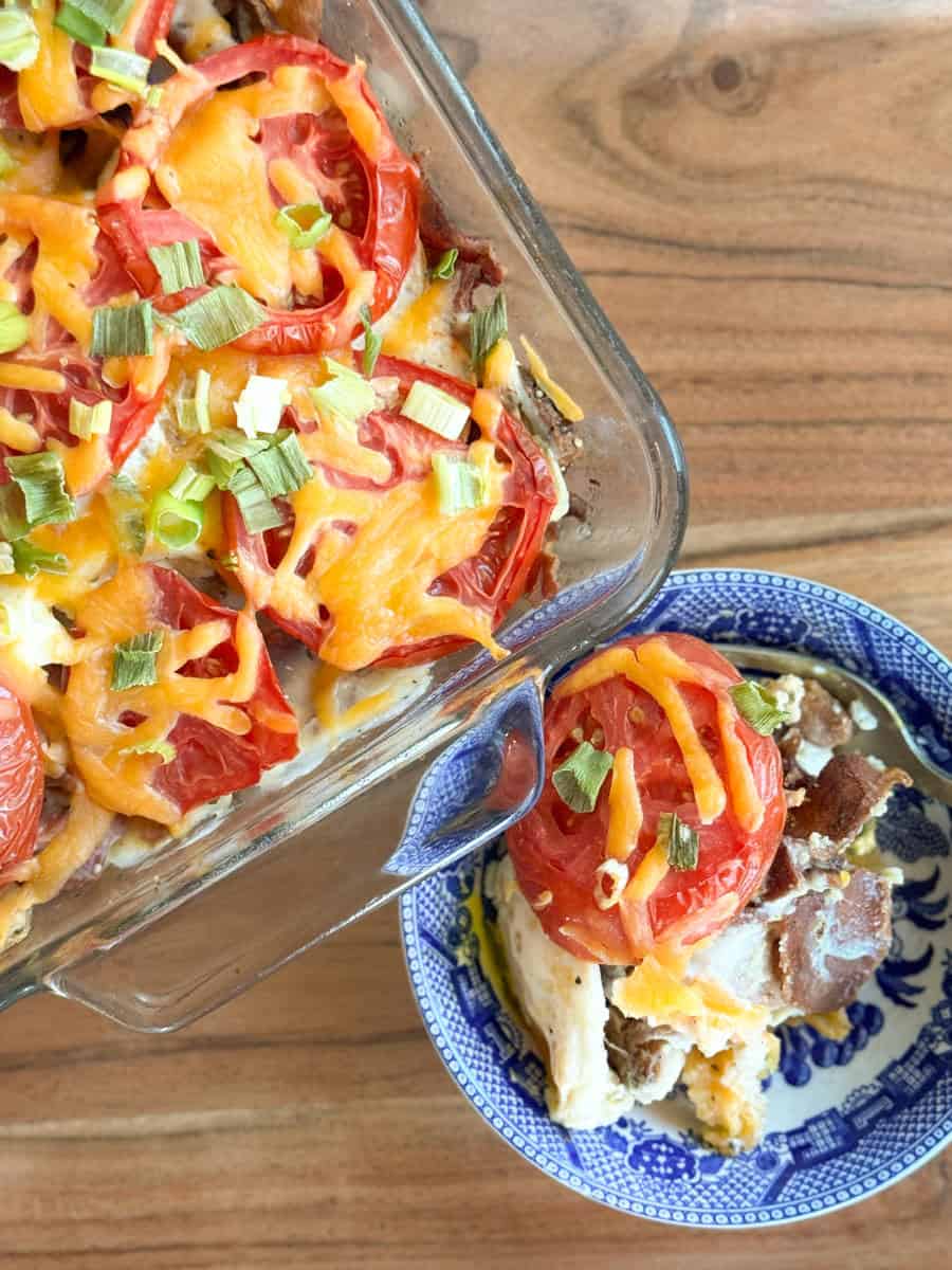 A glass baking dish of casserole topped with sliced tomatoes, melted cheddar cheese, and chopped green onions sits next to a blue-patterned bowl holding a serving of the casserole on a wooden surface.
