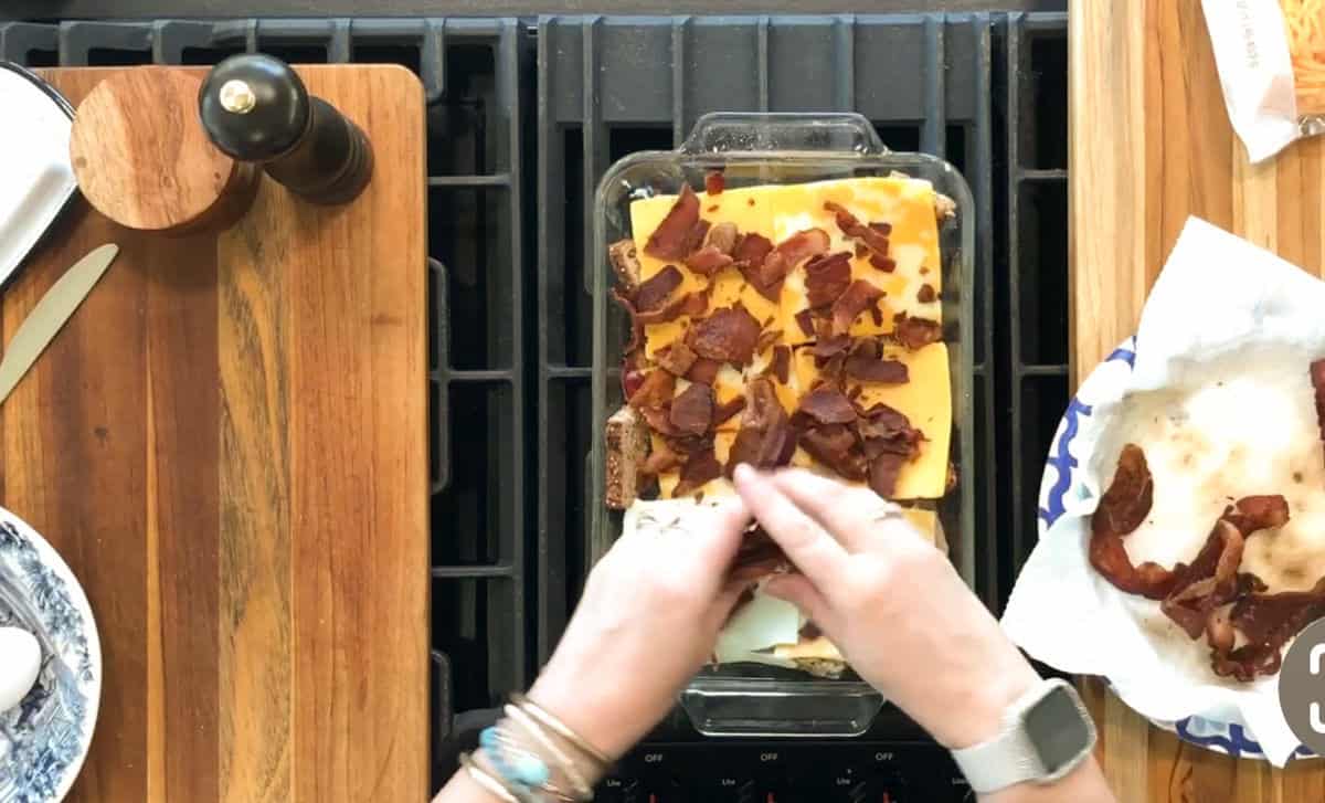 A person is assembling a dish in a glass baking pan on a stovetop, adding cooked bacon on top of cheese slices. A pepper grinder, knife, and paper towel with more bacon are on the wooden countertops nearby.