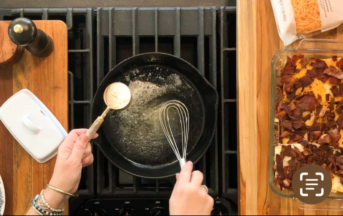 A person holds a whisk and a measuring spoon of flour over a cast iron skillet with melted butter on a stovetop; a pepper grinder, butter dish, and casserole are nearby.