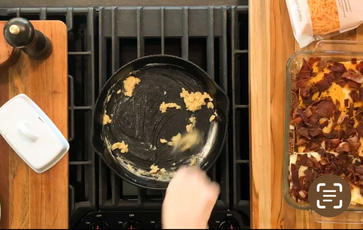 A person stirs a roux in a cast iron skillet on a stove. To the right, there is a glass baking dish with a casserole topped with pieces of cooked bacon. Various kitchen items are visible on the counter.