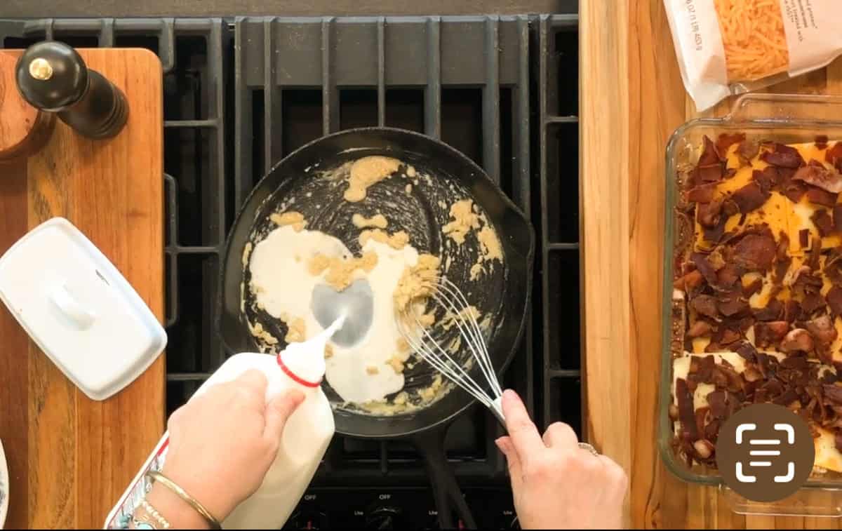 A person is pouring milk from a bottle into a skillet while stirring the contents with a spoon and a whisk. Ingredients, including a casserole topped with bacon, are on the counter nearby.