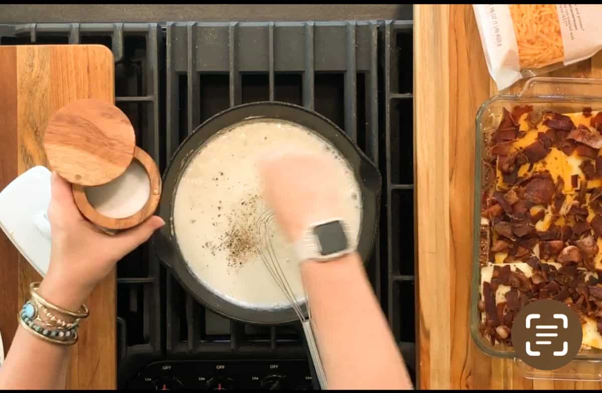 A person sprinkles seasoning into a saucepan of white sauce on the stove while stirring. Next to the stove is a dish of food topped with bacon, and an open bag of shredded cheese on a wooden surface.