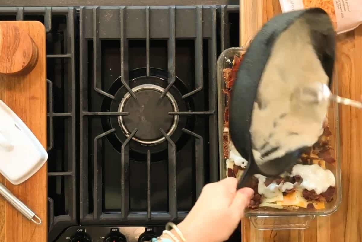 A person pours a white sauce from a pan onto a glass baking dish with layered ingredients on a wooden countertop beside a gas stove and kitchen utensils.