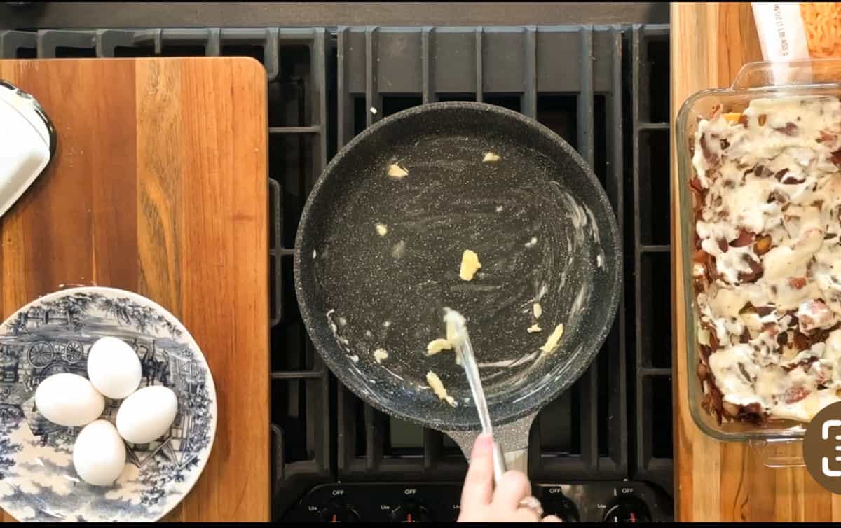 A hand holds tongs over a stovetop with a nonstick pan containing a few bits of food. To the left, a plate holds three eggs. To the right, there is a glass baking dish filled with a cooked casserole.