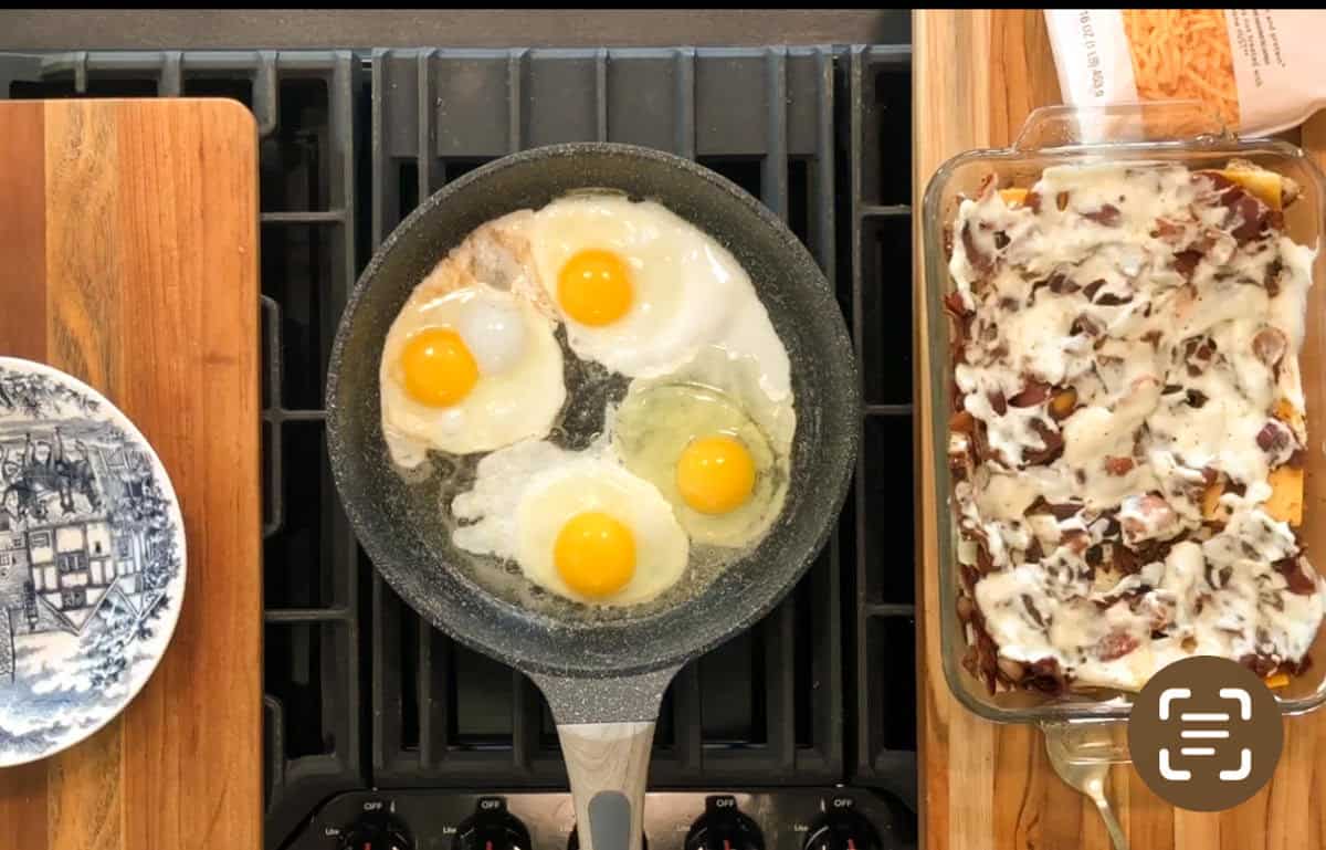 Four eggs are frying in a skillet on a stovetop. Next to the pan is a casserole dish filled with a baked dish topped with melted cheese and bacon pieces. A blue and white plate sits to the left on a wooden surface.