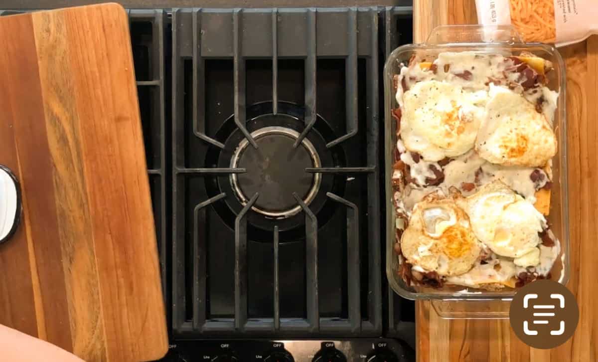 A glass baking dish containing casserole topped with four fried eggs sits on the right side of a stove, with a wooden cutting board and a person's left forearm visible on the left side of the image.