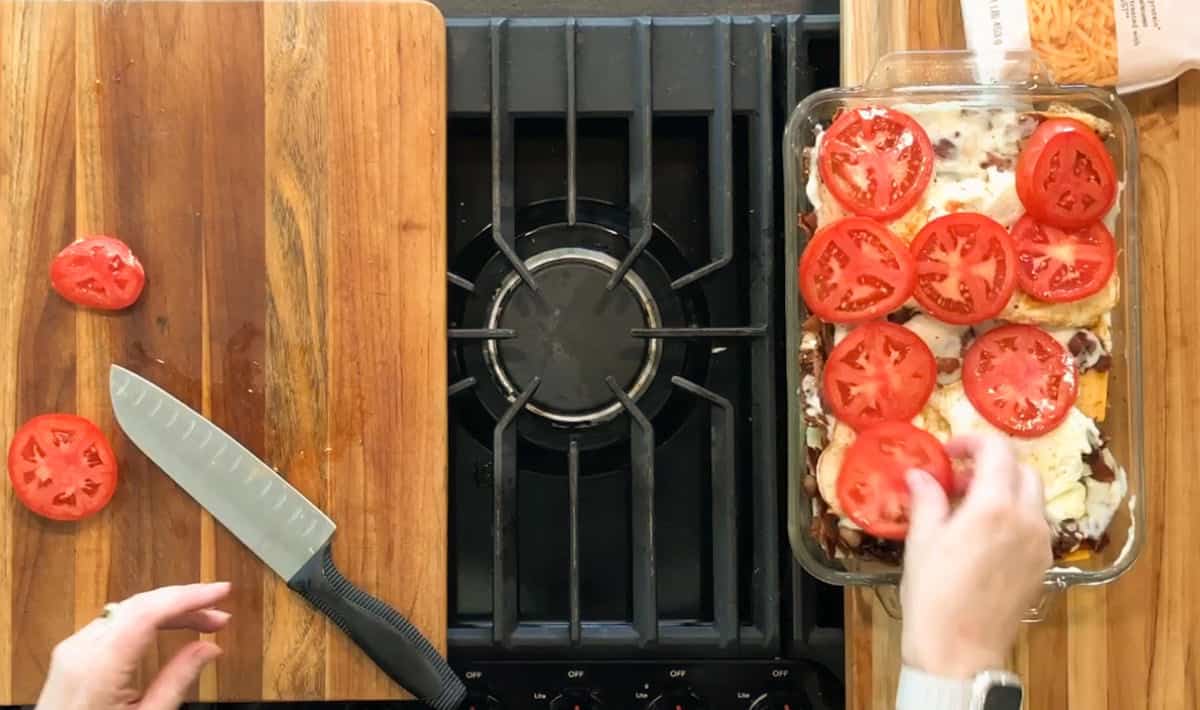 A person places fresh tomato slices on top of a casserole in a glass dish next to a stovetop. A cutting board with a knife and tomato slices is on the left side.
