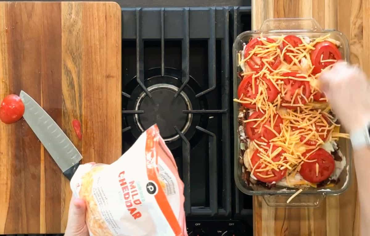 A hand sprinkles shredded mild cheddar cheese over a casserole topped with tomato slices. A cutting board with a knife and tomato is on the left, next to a stove with the casserole dish on the right.