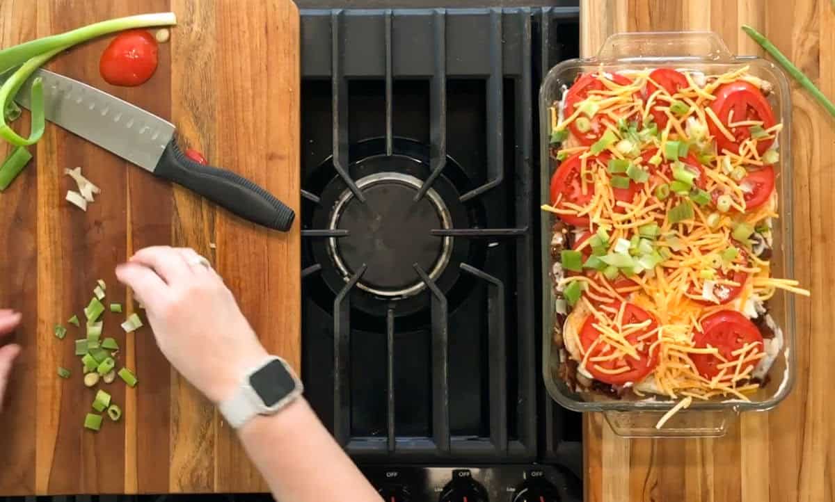 A person chops green onions on a wooden cutting board beside a stove. Next to the stove is a glass dish filled with a casserole topped with sliced tomatoes, shredded cheese, and chopped green onions.