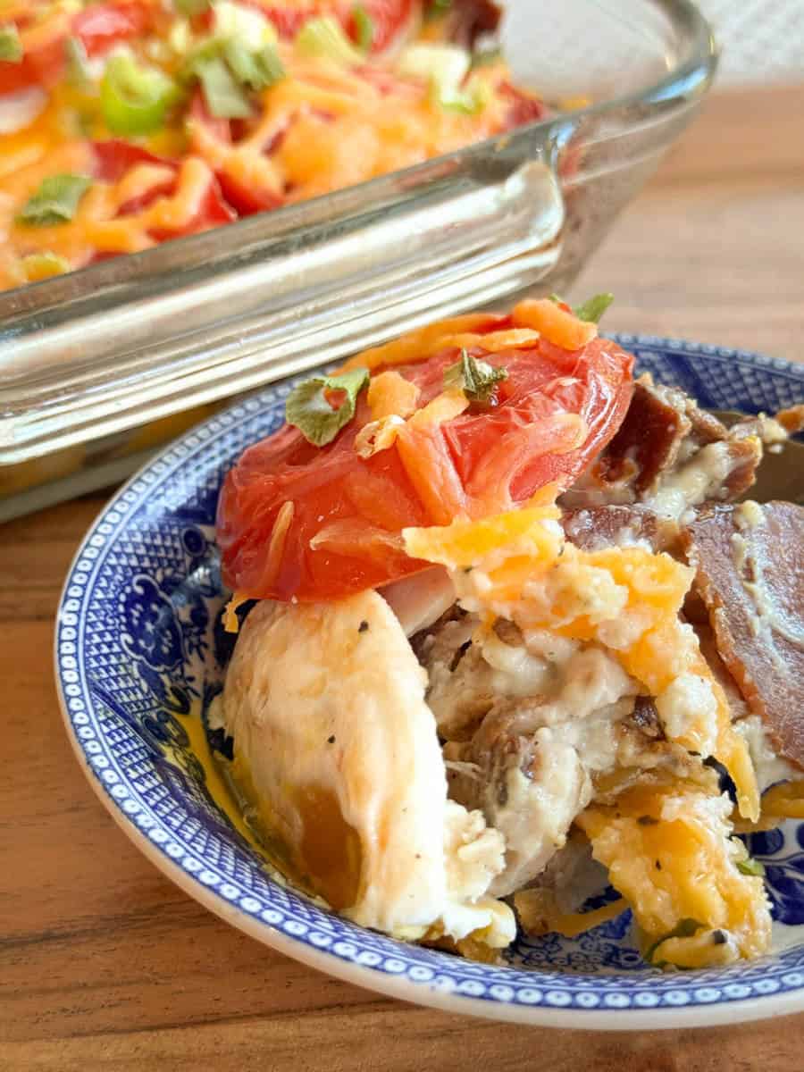 A blue and white bowl with a serving of baked chicken casserole topped with melted cheese, tomato, and herbs. A glass baking dish with more of the casserole is in the background on a wooden surface.