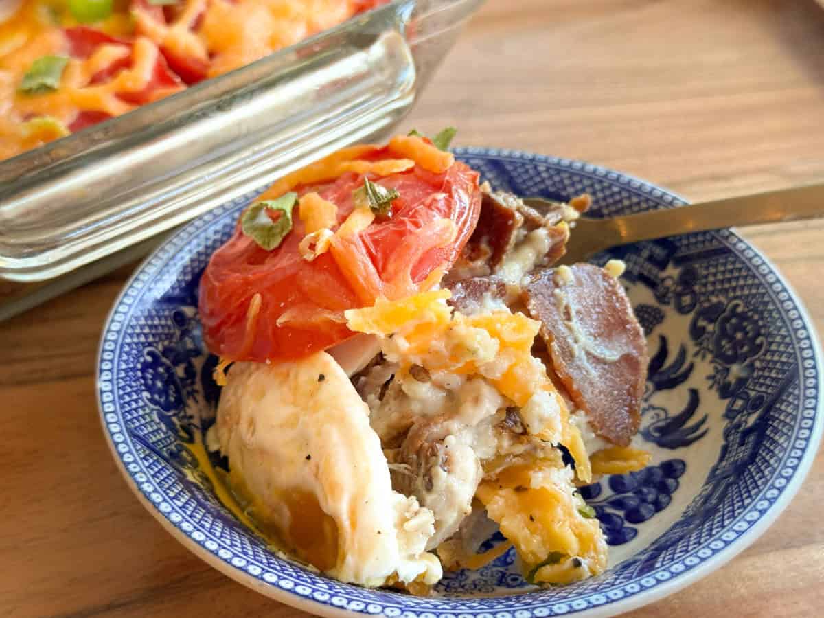 A close-up of a blue and white patterned bowl containing a serving of casserole with layers of baked eggs, cheese, bacon, and sliced tomatoes. A fork is placed in the bowl, and a casserole dish is in the background.