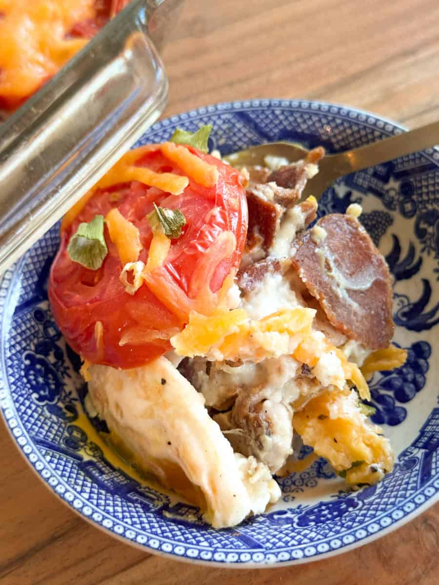 A serving of a baked casserole with sliced tomatoes, potatoes, cheese, and herbs is shown on a blue and white patterned plate with a fork, next to a glass baking dish on a wooden surface.