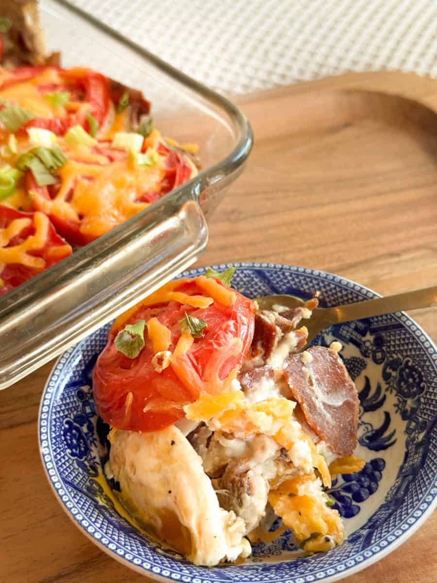 A serving of baked casserole topped with sliced tomatoes, cheese, and chopped herbs is in a blue and white bowl beside a glass baking dish with the remaining casserole on a wooden surface.