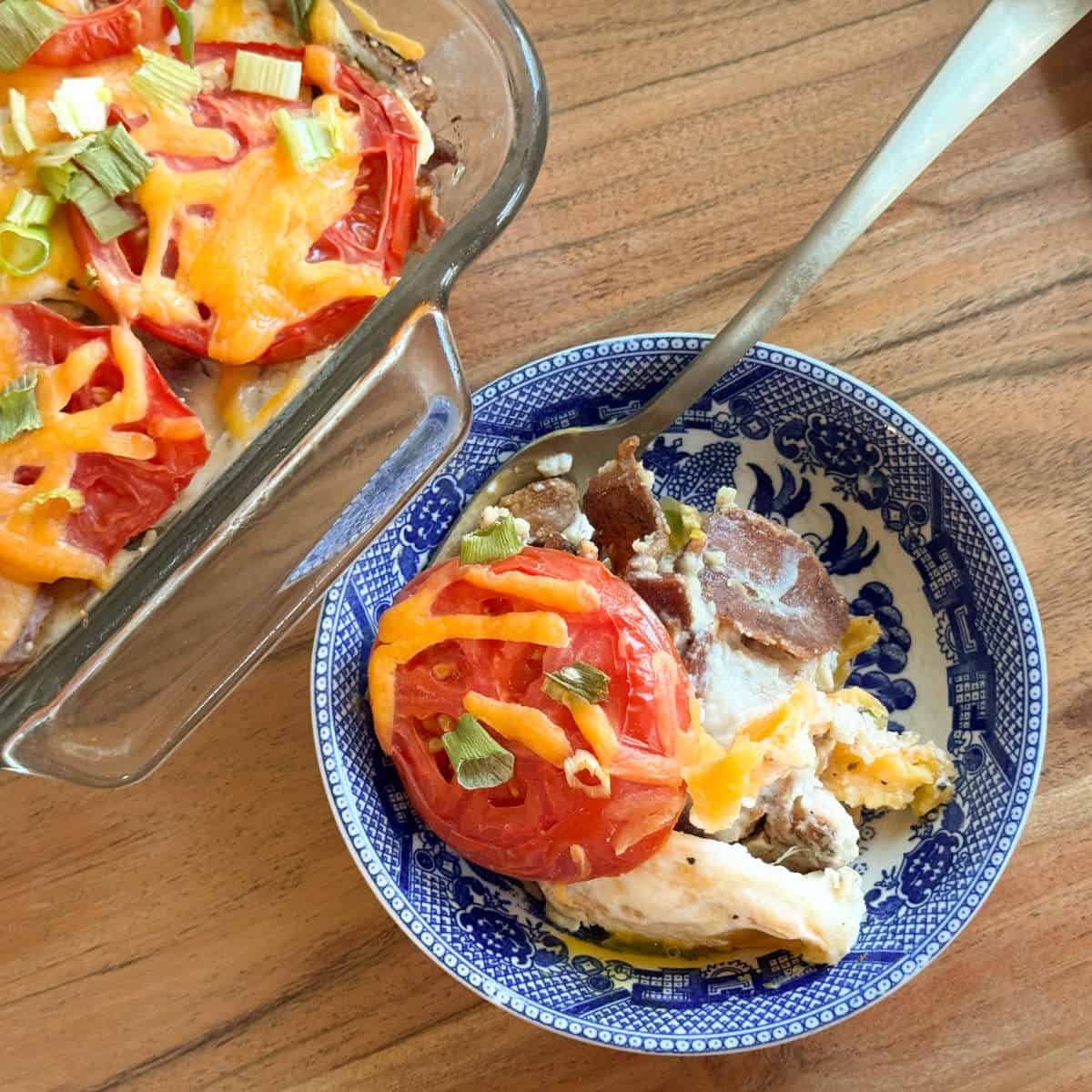 A serving of casserole with tomato slices, melted cheddar cheese, green onions, and visible potato slices is plated in a blue-and-white bowl next to the main casserole dish on a wooden table.