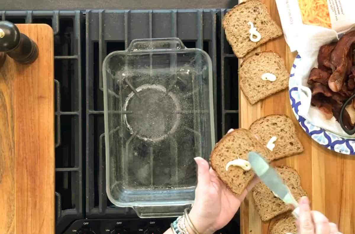 A person spreads mayonnaise on slices of whole wheat bread next to a glass baking dish on a stovetop, with cooked bacon and shredded cheese visible nearby.