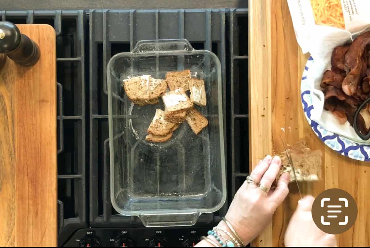 A person chops bread into cubes on a wooden cutting board next to a glass baking dish containing more bread cubes, with a plate of cooked bacon nearby on the right. The setup is on a stovetop and wooden countertop.