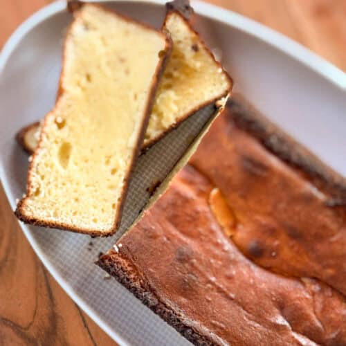 A close-up of a rectangular loaf cake on a white plate, with two slices cut and laid on top. The cake has a golden brown crust and a light yellow, dense interior. The plate rests on a wooden surface.