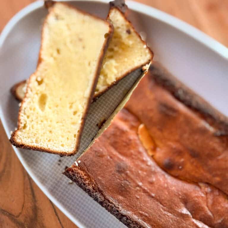 A close-up of a rectangular loaf cake on a white plate, with two slices cut and laid on top. The cake has a golden brown crust and a light yellow, dense interior. The plate rests on a wooden surface.