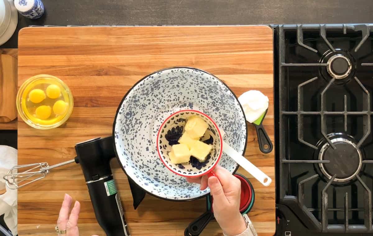 A person holds a small bowl of butter over a large speckled mixing bowl on a wooden cutting board. Nearby are eggs in a glass bowl, a hand mixer, measuring cups, and a stovetop.