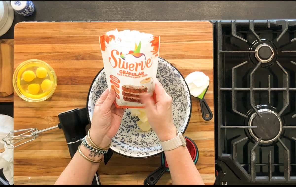 A person holds a bag of Swerve Granular sweetener over a mixing bowl with butter. Nearby are eggs in a glass bowl, measuring cups, an electric mixer, and a stovetop on a wooden counter.