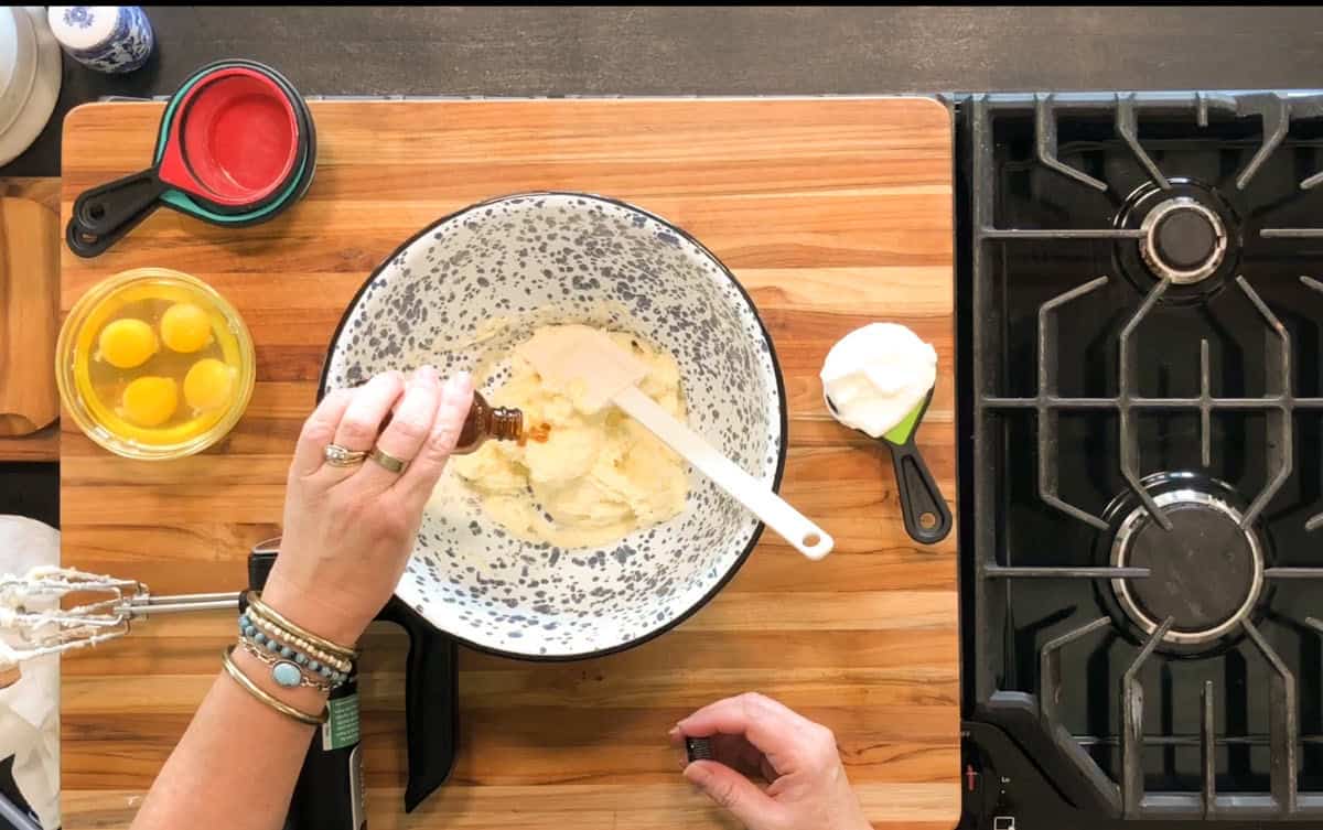 A person adds liquid from a small bottle into a mixing bowl with batter on a wooden countertop, next to eggs in a bowl, sour cream, and a gas stovetop. A hand mixer and spatula are also visible.