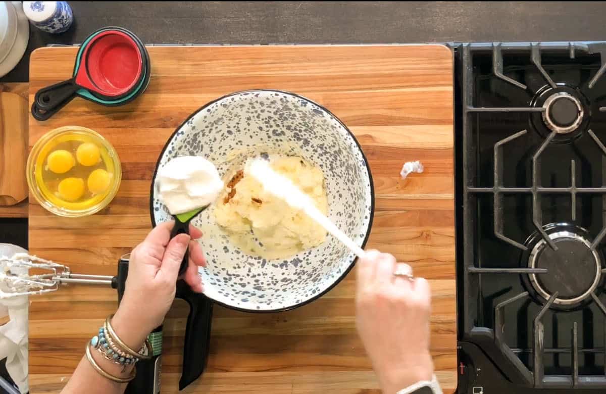 A person adds a cup of sour cream to a speckled mixing bowl containing other ingredients. Three eggs in a glass bowl, a hand mixer, and measuring cups are on a wooden countertop beside a stovetop.