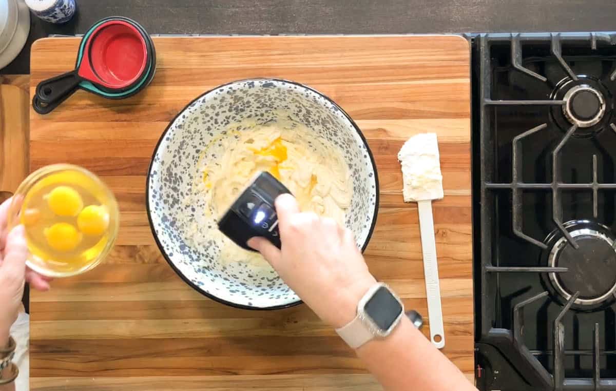 A person is beating a mixture in a speckled mixing bowl with a hand mixer on a wooden cutting board. Another hand holds a small bowl with three eggs. A spatula and measuring cups are nearby beside a stove.