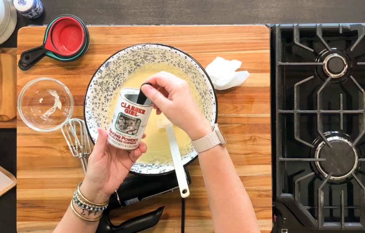 A person adds Clabber Girl baking powder to a mixing bowl with a yellow batter on a wooden countertop next to a gas stove, handheld mixer, measuring cups, and a spatula.