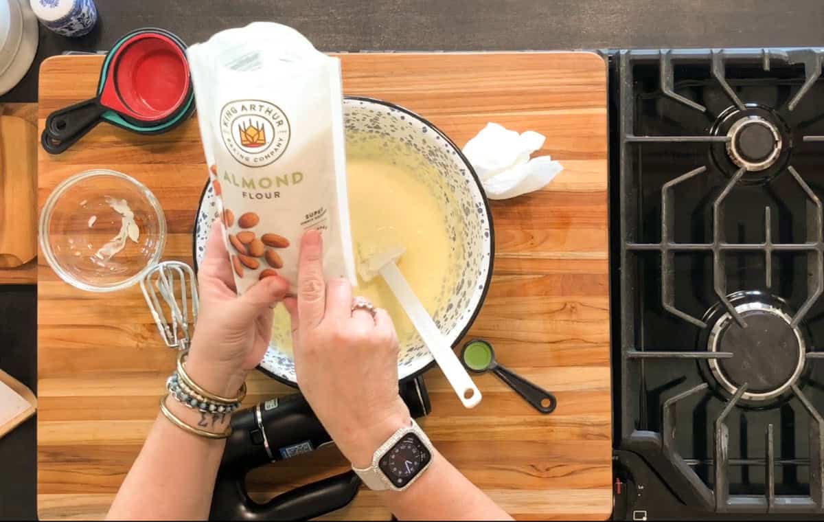 A person holding a bag of almond flour above a mixing bowl with batter on a wooden countertop, next to a glass bowl, measuring spoons, and a stove.