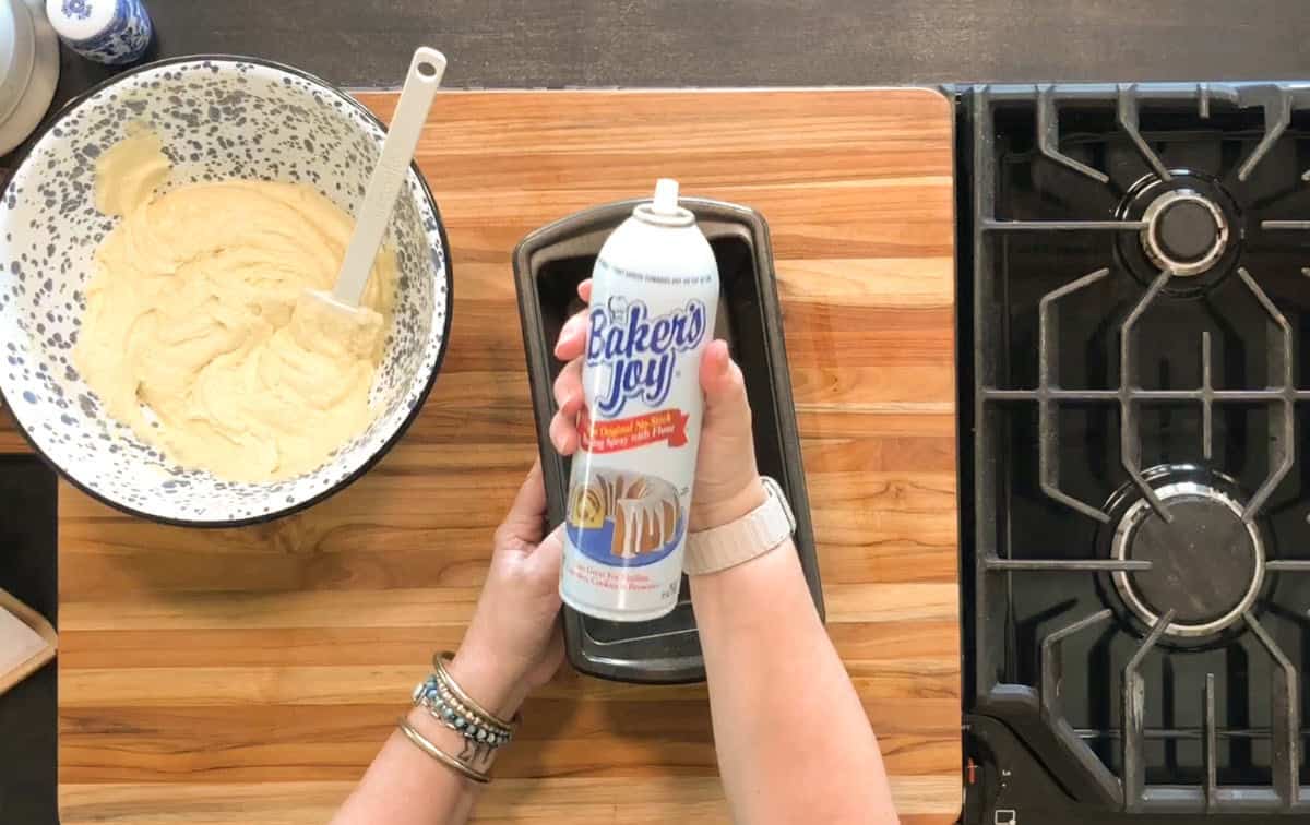 A person holds a can of Baker's Joy nonstick spray over a loaf pan on a wooden cutting board, with a bowl of batter and a spatula nearby, next to a stove.