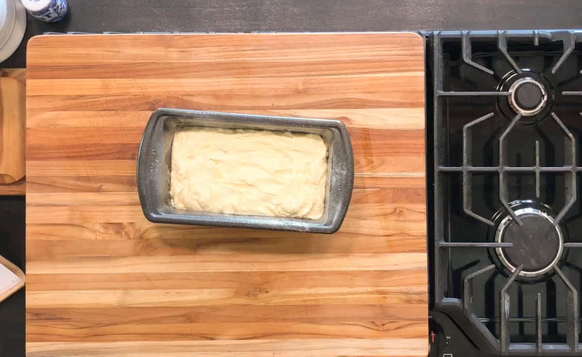 A metal loaf pan filled with unbaked batter sits on a wooden cutting board next to a black gas stove on a dark kitchen countertop.