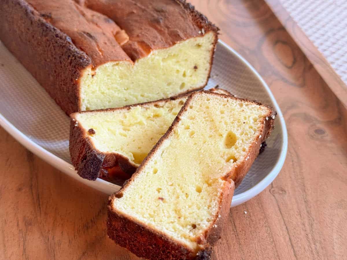 A loaf of pound cake with a golden brown crust is sliced, revealing a dense, yellow interior. Two slices are lying flat on a white plate on a wooden table.