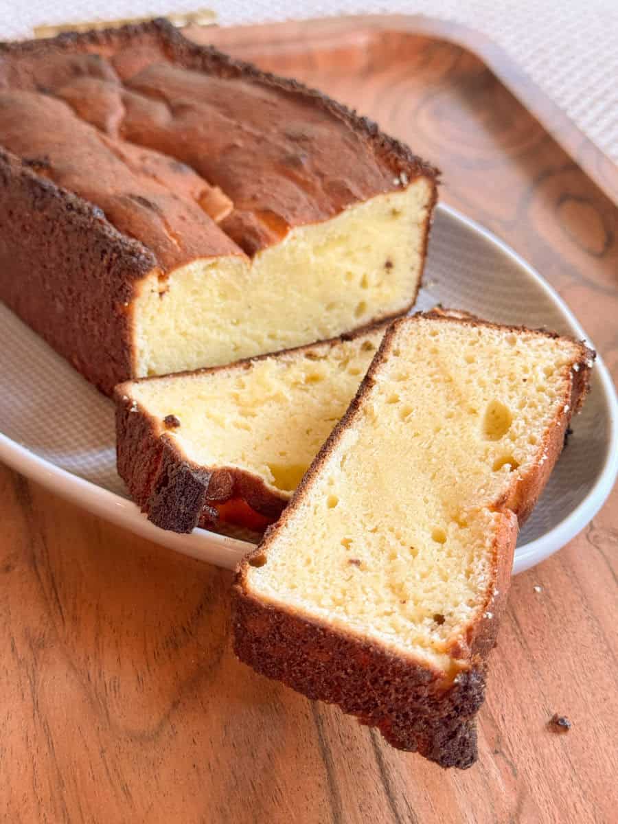 A loaf of golden yellow pound cake with a dark brown crust sits on a plate. Two slices are cut and laid in front of the loaf. The cake has a dense, moist texture and is placed on a wooden surface.