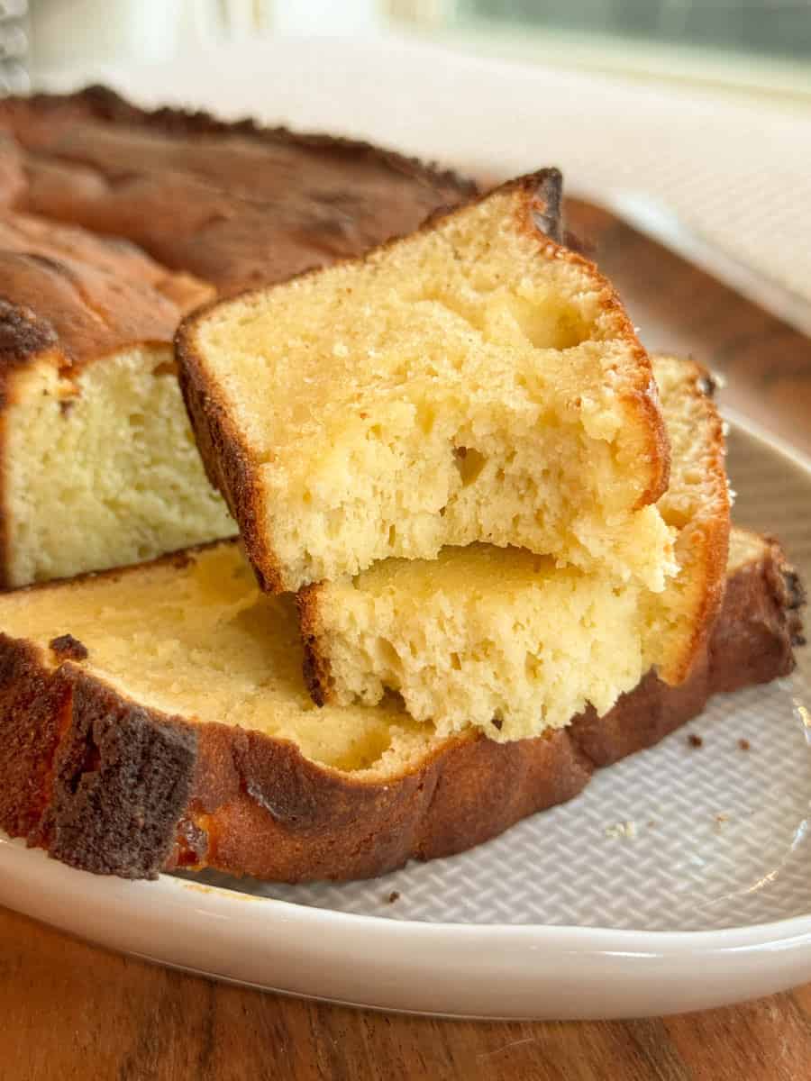 Close-up of several slices of golden-yellow pound cake with a slightly browned crust, stacked on a white textured plate. The cake appears moist and dense with a soft, spongy interior.