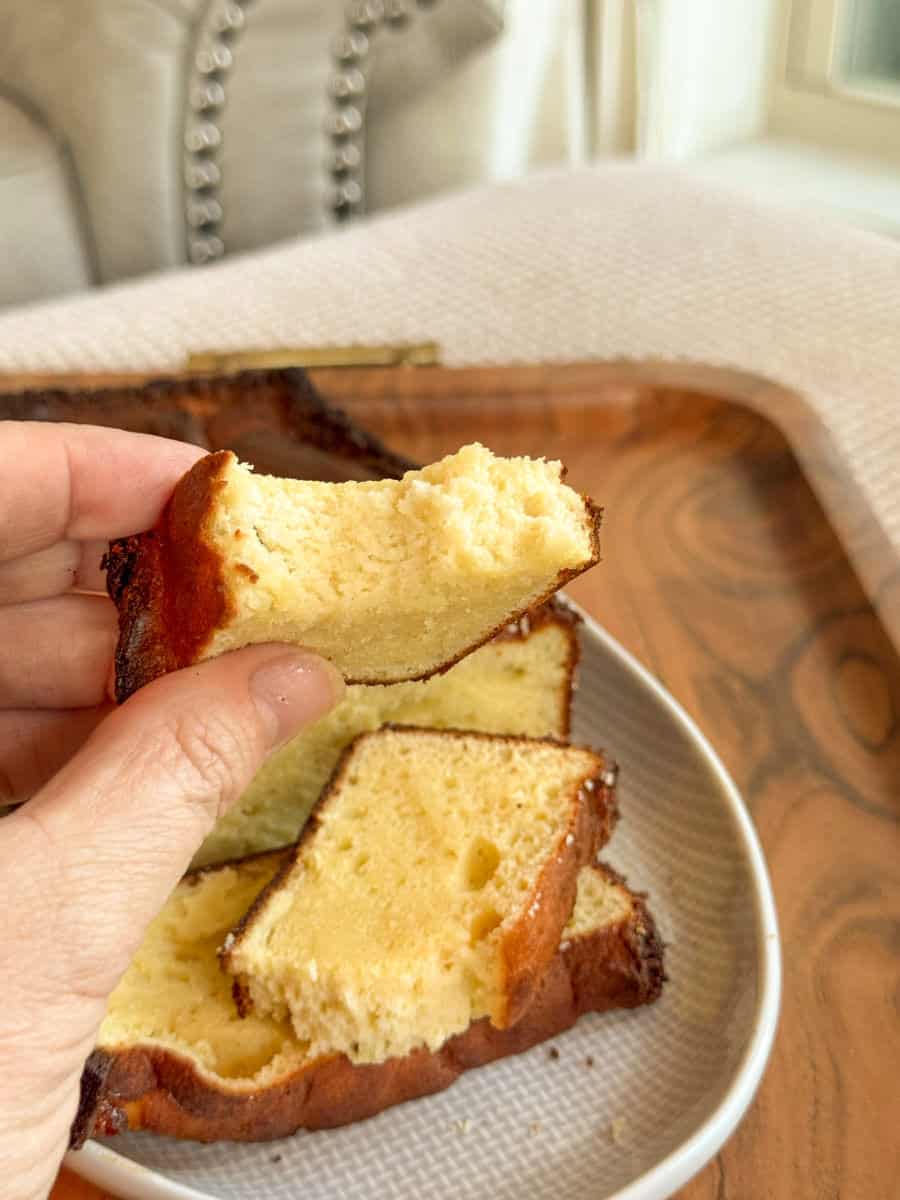 A hand holds up a thick slice of yellow pound cake with a bite taken out of it. Several more slices of the same cake are arranged on a white plate on a wooden tray in the background.