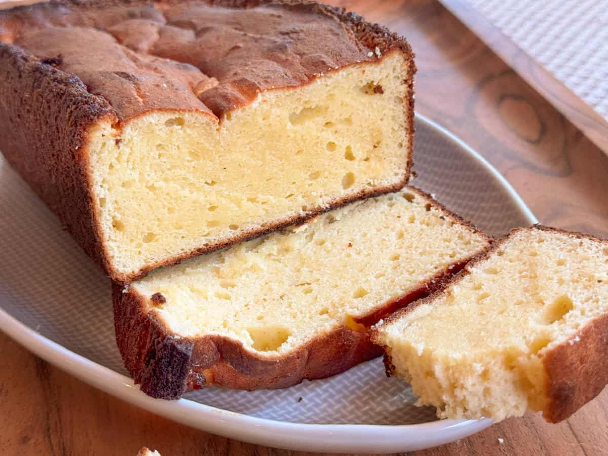 A loaf of plain pound cake with a golden-brown crust is shown on a plate. Two slices have been cut, with one partially separated from the rest of the loaf. The cake has a dense, moist texture.