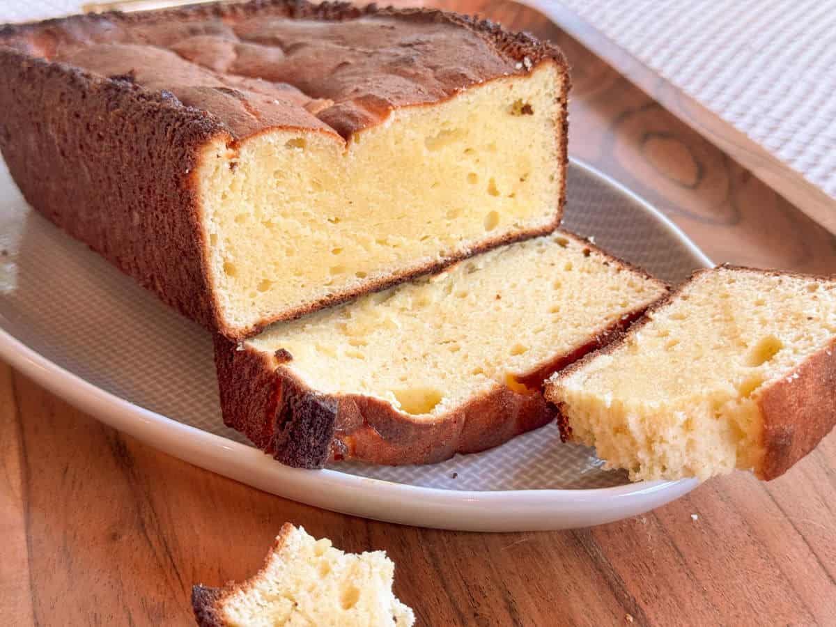 A loaf of pound cake sits on a white plate atop a wooden surface. Two slices have been cut from the loaf, with one slice partially separated and a small crumb in the foreground.