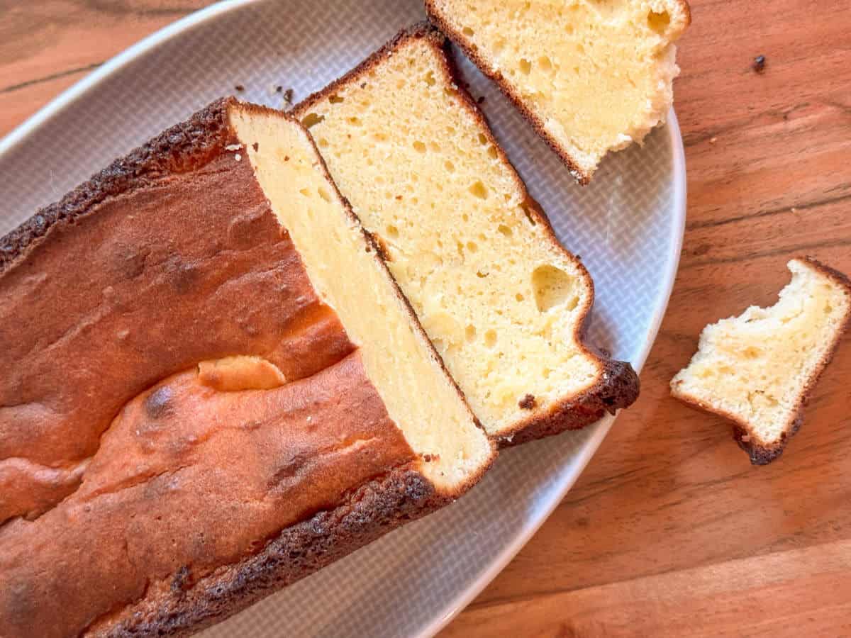 A rectangular pound cake with a golden-brown crust is shown on a white plate. Two slices have been cut from the loaf, with one small piece broken off and placed beside the slices on a wooden surface.