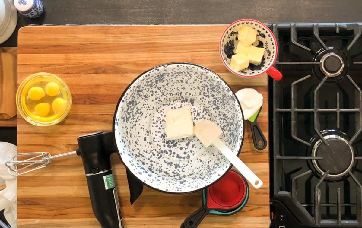 A mixing bowl with butter and a spatula sits on a wooden countertop next to eggs in a glass bowl, a mixer, measuring cups, and a gas stove. Butter and cream cheese are in a small bowl nearby.