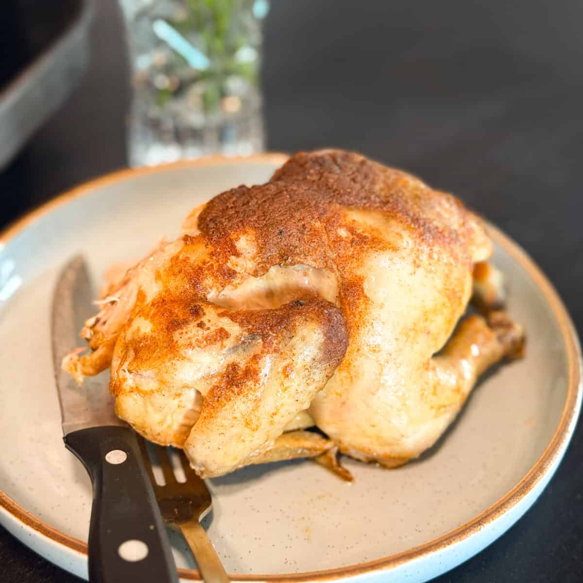 A whole roasted chicken with browned skin sits on a round plate next to a knife and fork. The background includes a glass container with herbs, slightly out of focus.