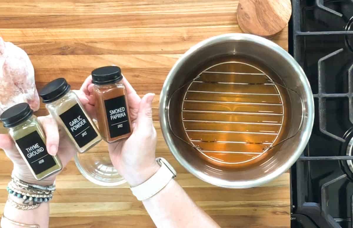 A person holds three spice jars labeled thyme ground, garlic powder, and smoked paprika over a wooden counter next to a pot with liquid and a metal rack on a stove.