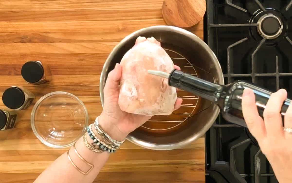 A person pours oil from a bottle onto a raw chicken breast held over a metal bowl on a wooden countertop near spice jars and a stove.