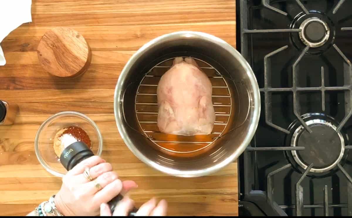A person grinds pepper into a bowl of spices next to a large piece of meat resting on a rack inside an Instant Pot on a wooden countertop beside a stovetop.