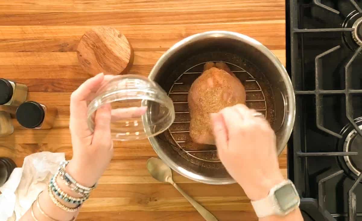 A person is seasoning a piece of meat in a metal pot on a wooden countertop. There are spice containers, a small wooden lid, a spoon, and a gas stove visible in the background.